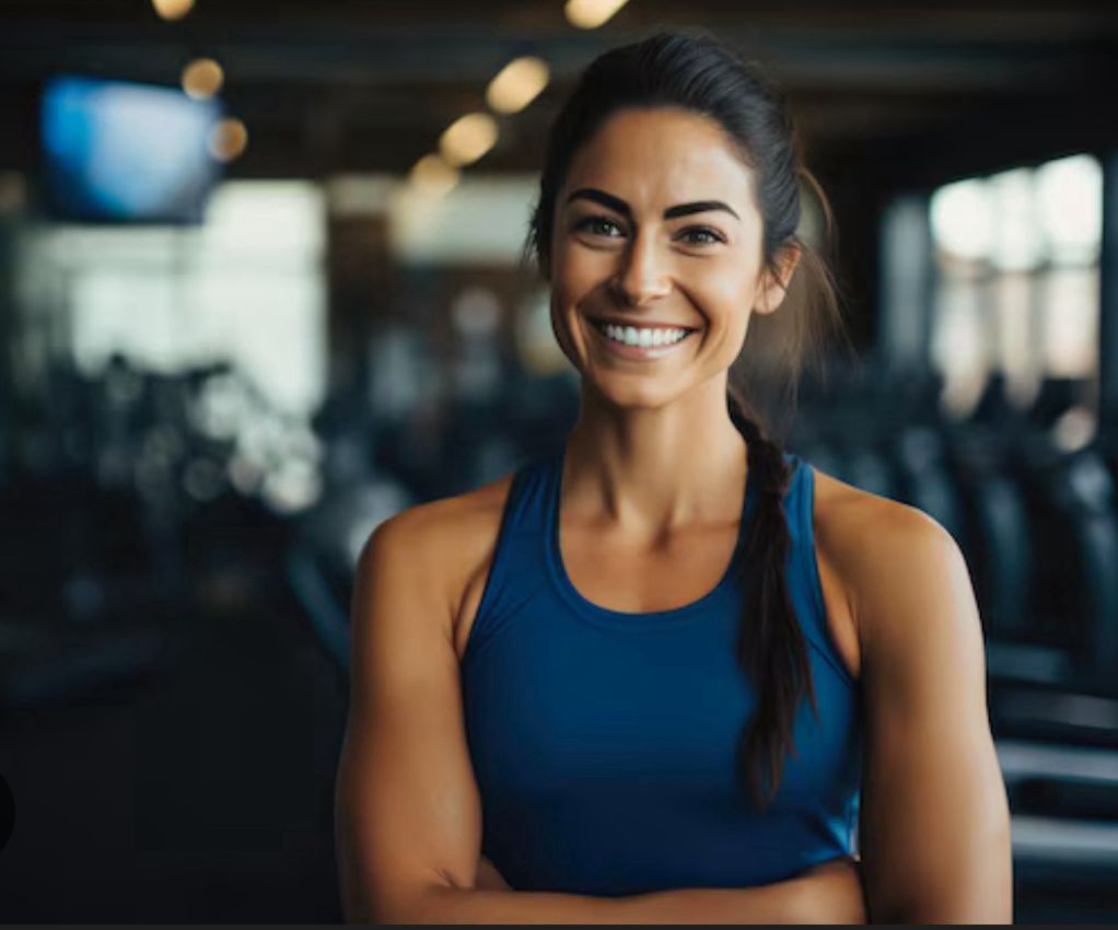 Smiling woman in a blue athletic tank top standing in a gym setting with fitness equipment in the background.